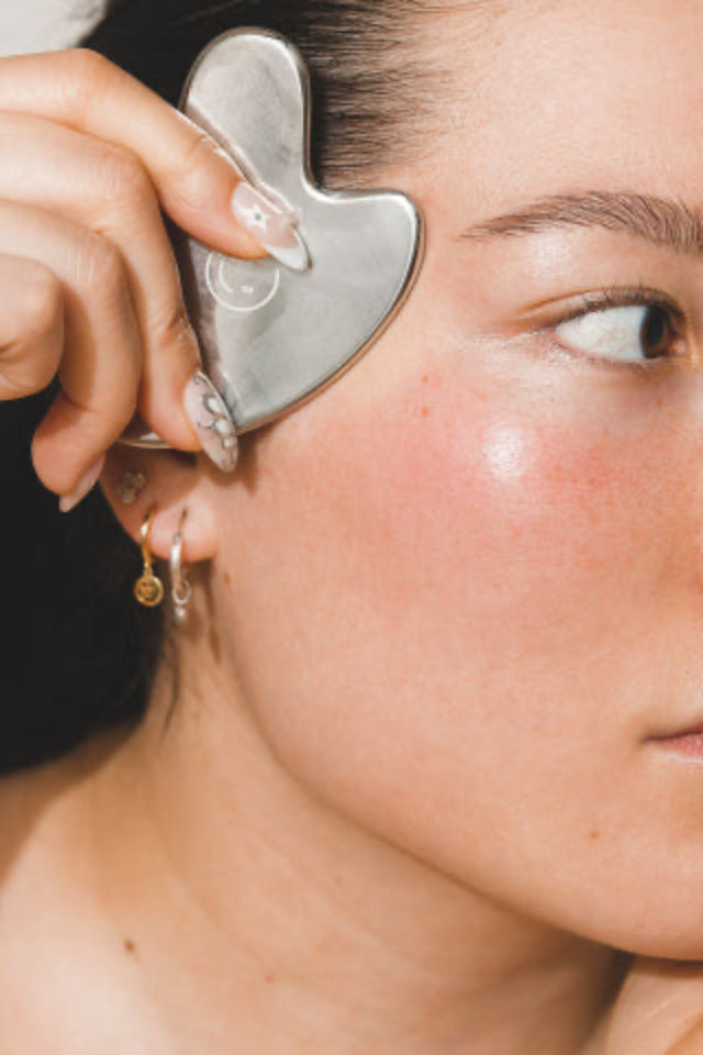 Close-up of a person using a stainless steel gua sha tool on their cheekbone, highlighting smooth, radiant skin and minimalist jewelry; part of the skincare ritual from Kid Sister Skincare.