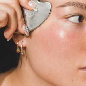 Close-up of a person using a stainless steel gua sha tool on their cheekbone, highlighting smooth, radiant skin and minimalist jewelry; part of the skincare ritual from Kid Sister Skincare.